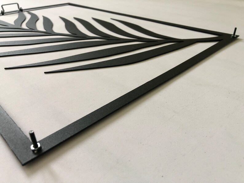 Puede incluir: Arte de pared de metal negro con un dise&ntilde;o estilizado de hoja de palma. El arte est&aacute; enmarcado con un marco de metal negro y tiene dos tornillos en la parte inferior.