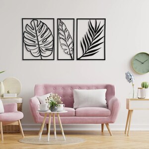Puede incluir: Tres piezas de arte de pared de metal negro con siluetas recortadas de hojas tropicales. Las piezas est&aacute;n dispuestas en una fila en una pared blanca sobre un sof&aacute; rosa y un sill&oacute;n rosa.