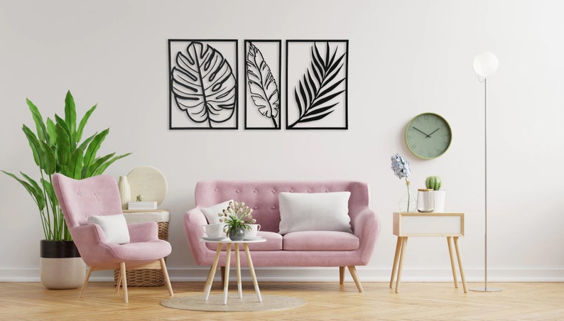 Puede incluir: Tres piezas de arte de pared de metal negro con siluetas recortadas de hojas tropicales. Las piezas est&aacute;n dispuestas en una fila en una pared blanca sobre un sof&aacute; rosa y un sill&oacute;n rosa.
