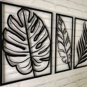 Puede incluir: Tres piezas de decoraci&oacute;n de pared de metal negro con dise&ntilde;os recortados de hojas tropicales. Las piezas son rectangulares y tienen un estilo simple y moderno.
