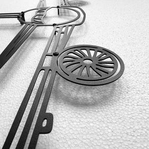 May include: Black metal wall art featuring an abstract design with geometric shapes and a circular element resembling a wheel. The artwork is mounted on a textured white surface, creating a contrast in tones.