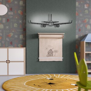 May include: A black metal airplane wall art with a light behind it is mounted above a roll of paper with a house drawing. The room has a yellow rug with a lion face design and a gray patterned wallpaper.