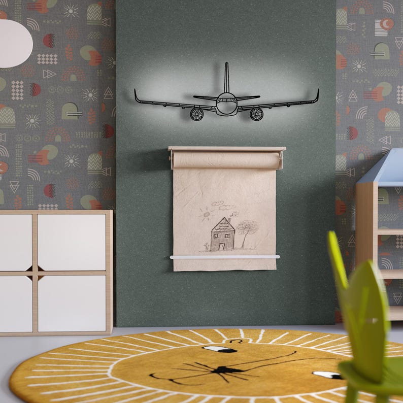 May include: A black metal airplane wall art with a light behind it is mounted above a roll of paper with a house drawing. The room has a yellow rug with a lion face design and a gray patterned wallpaper.