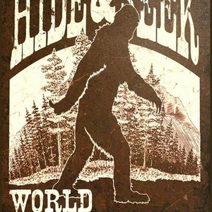 May include: A brown and white graphic design with a silhouette of Bigfoot walking through a forest. The text "HIDE & SEEK WORLD CHAMPION" is written in a vintage style font.