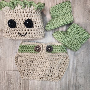 Puede incluir: Un conjunto de bebé tejido a crochet inspirado en el personaje Groot de las películas Guardianes de la Galaxie. El conjunto incluye un gorro, una cubierta de pañal y botines. El gorro tiene una parte superior verde con hojas y una cara marrón con ojos negros. La cubierta de pañal tiene una banda verde con dos botones marrones. Los botines son verdes y tienen una textura de crochet.