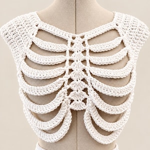 May include: A white crochet top featuring a structural design that mimics the appearance of a human rib cage. The garment is constructed with thick yarn, showcasing a central vertical spine pattern with curved, rib-like segments extending outward.