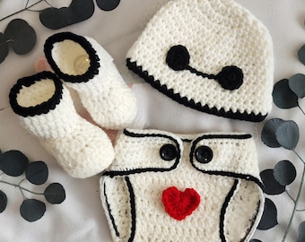May include: A white crocheted baby outfit with a black trim. The outfit includes a hat, diaper cover, and booties. The hat has a black eye design and the diaper cover has a red heart.