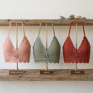May include: Three handmade crochet tops in warm blush, sage, and rust colors. Each top has a halter neck and a scalloped edge. The tops are displayed on gold stands with labels that read "Warm Blush", "Sage", and "Rust".