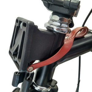 Op de afbeelding: Close-up van een zwart fietsstuur met een zwarte plastic beugel en een rode leren riem. De beugel is met een zilveren schroef aan het stuur bevestigd. De leren riem is gestikt met wit garen.