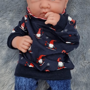 May include: A baby doll dressed in a navy blue sweatshirt featuring skiing gnomes and snowflakes, and blue denim jeans. The doll is lying on a grey, fluffy surface. The doll's height is approximately 30 cm.