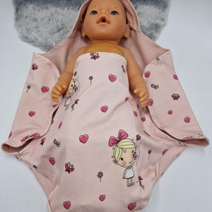 Könnte beinhalten: Eine Babypuppe, eingewickelt in eine rosa Kapuzen-Decke. Die Decke ist mit rosa Herzen, Schmetterlingen, Blumen und einem Cartoon-Mädchen verziert. Die Puppe hat blaue Augen und liegt auf einer grauen, flauschigen Oberfläche.