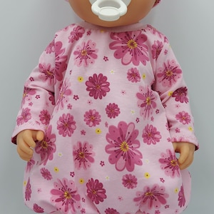 以下が含まれることがあります： ピンクのベビー人形は、ピンクの花柄のドレスとピンクのヘッドバンドを着て、口に白いおしゃぶりをしています。