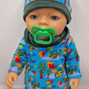 May include: A doll wearing a blue beanie and a long-sleeved shirt, both featuring a colorful dragon and tree pattern. The doll has a dark green collar, dark green pants, and a bright green pacifier in its mouth. The doll has blue eyes.