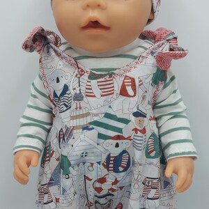 以下が含まれることがあります： 白いと緑のストライプの長袖シャツ、模様入りのジャンプスーツ、お揃いの帽子など、航海をテーマにした衣装を着た人形。ジャンプスーツには、コアラ、ヨット、赤と青のアクセントが施されています。