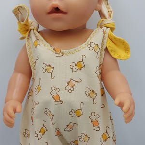 以下が含まれることがあります： クリーム色のジャンプスーツを着たベビー人形。ジャンプスーツには、黄色いマウスのプリントが施されています。ジャンプスーツは、肩で結ぶことができる黄色のストラップが付いています。