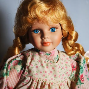 May include: A porcelain doll with blonde, curly hair and bright blue eyes. The doll wears a pink floral dress with a high collar. The doll's face is painted with rosy cheeks and a gentle smile. A classic collectible doll.