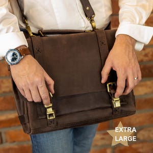 A person is holding a brown leather satchel with a strap across their body, wearing a watch on their left wrist and a ring on their right hand.