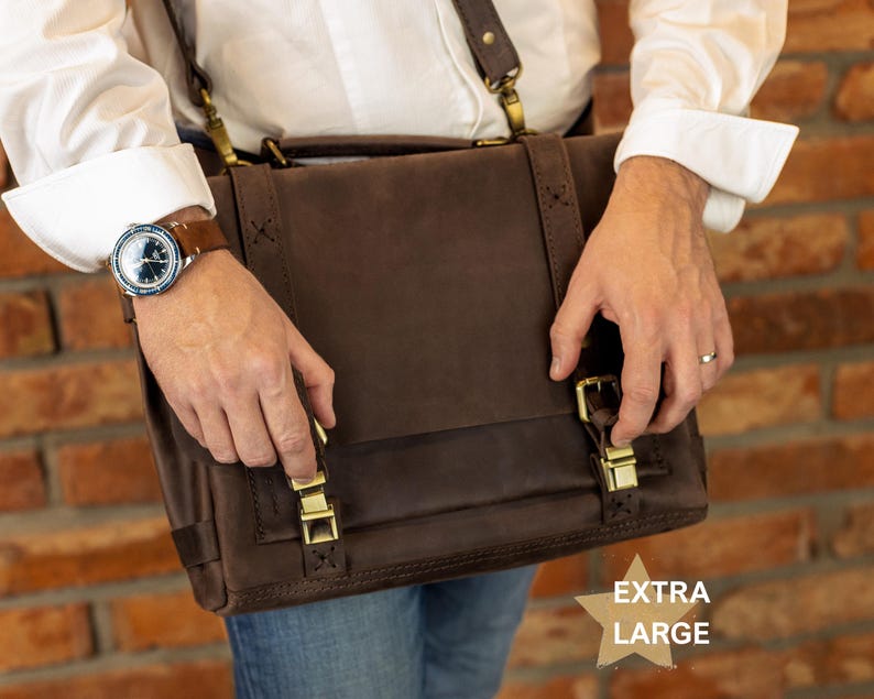 A person is holding a brown leather satchel with a strap across their body, wearing a watch on their left wrist and a ring on their right hand.