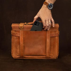 A hand is holding a black iPhone out of a brown leather briefcase or satchel.