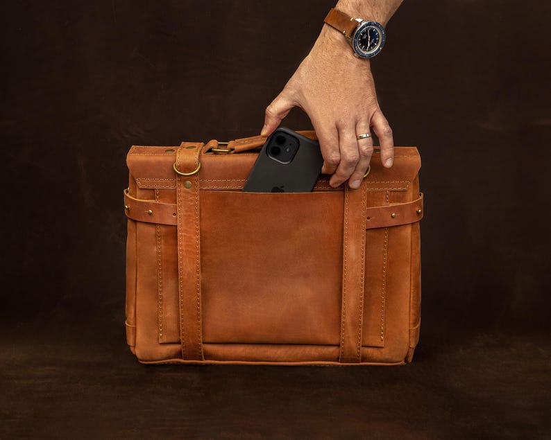 A hand is holding a black iPhone out of a brown leather briefcase or satchel.
