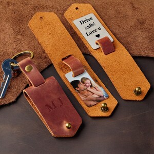 May include: A brown leather keychain with a personalised inscription "MJ" and a silver metal tag with a photo of a couple. The keychain also includes a silver metal tag with the text "Drive safe! Love u" and a heart symbol.