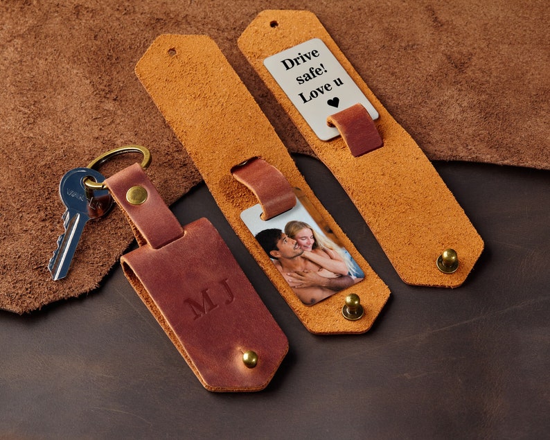 May include: A brown leather keychain with a personalised inscription "MJ" and a silver metal tag with a photo of a couple. The keychain also includes a silver metal tag with the text "Drive safe! Love u" and a heart symbol.