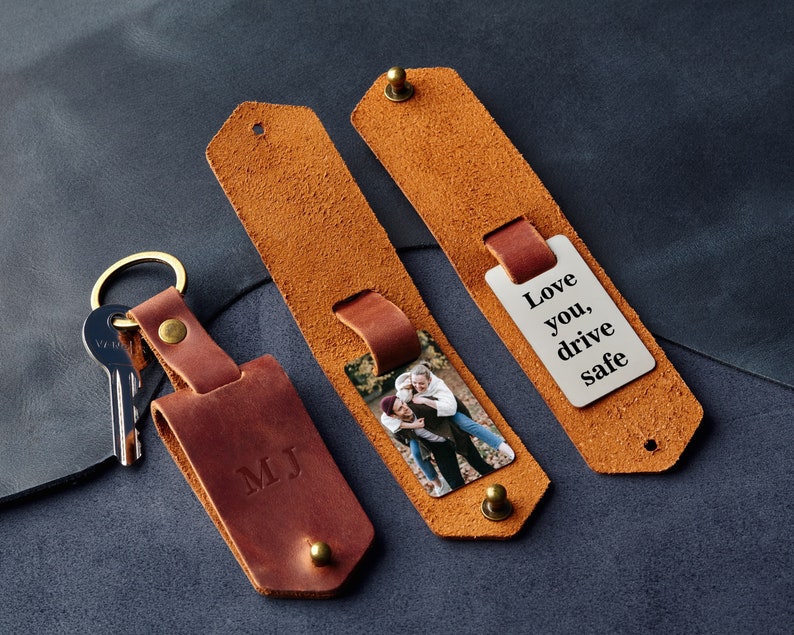 May include: A brown leather keychain with a silver tag that says "Love you, drive safe". The keychain also has a photo insert and a personalised initial tag that says "MJ".