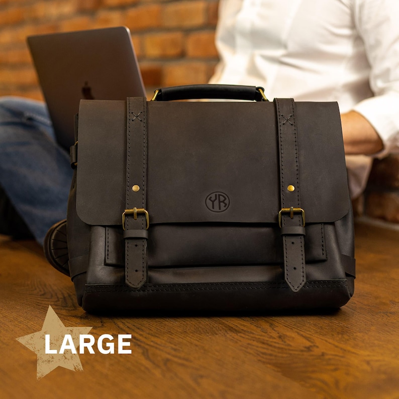 A large black leather satchel with a laptop bag is placed on a wooden surface, with a person's leg visible in the background.