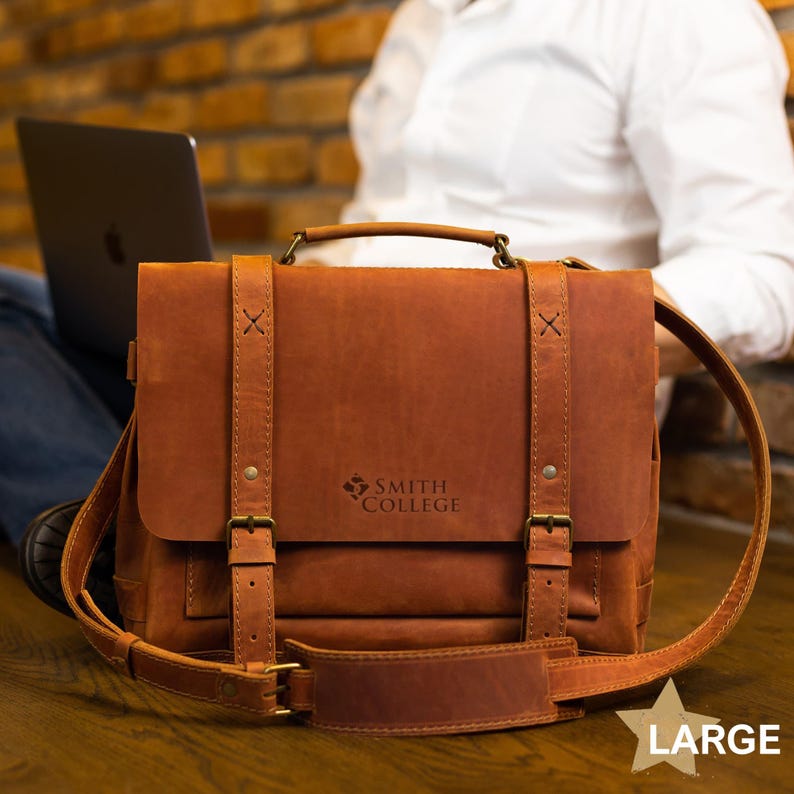 A brown leather satchel with the Smith College logo, placed on a wooden surface, with a laptop and a person's arm visible in the background.