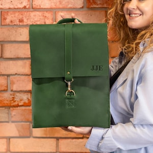 May include: A green leather backpack with a top handle and a flap closure. The bag features a strap with a gold-colored clasp and the initials "JJE" embossed on the front. The bag is held in front of a brick wall.