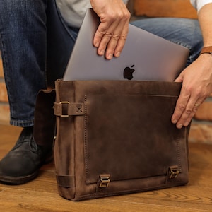 May include: A brown leather messenger bag with a flap closure and brass-colored hardware. A silver laptop with the Apple logo is being placed inside the bag. The bag is made of a distressed leather material.
