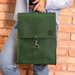 May include: A dark green leather backpack with a gold buckle and a monogrammed circle with the letters "SGA" on the front.