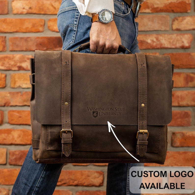 A person is holding a brown leather satchel with a Washington State University logo on it, standing in front of a brick wall.