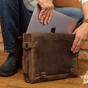 A person is holding a laptop in a brown leather bag, with a brick wall in the background.