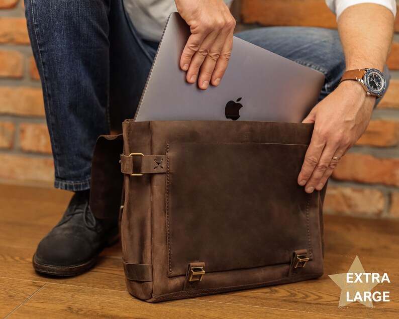 A person is holding a laptop in a brown leather bag, with a brick wall in the background.