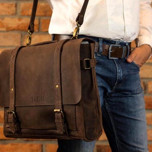 May include: A dark brown leather messenger bag featuring two vertical straps with brass-toned buckles, a top handle, and an adjustable shoulder strap. The front flap is embossed with the letters HEB and secured with metal hardware.