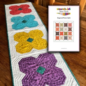 May include: A white table runner with a teal border and four colorful flower designs. The flowers are red, blue, yellow, and purple. The runner is quilted with a white background and a pattern of small circles in the purple flower. The runner is on a wooden table with a pattern of a quilt design on a white paper with the text "Sew York Dogwood Flower Quilt www.sewyork.co.uk".