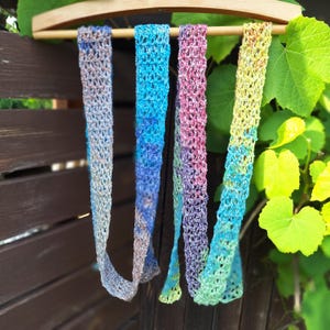 Puede incluir: Cuatro bufandas coloridas de tejido abierto cuelgan de una barra de madera. Las bufandas muestran un degradado de colores, incluyendo azul, morado, rosa y amarillo. Las bufandas se exhiben contra una valla de madera y follaje verde.