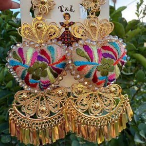 May include: Gold-toned filigree earrings with colorful embroidered heart-shaped centers. The earrings feature a floral design at the top and a fringe at the bottom. The text "T&L" is visible on the earring card.