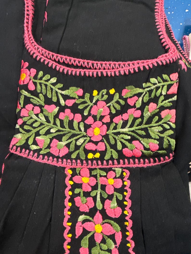 May include: Black garment with pink floral embroidery and green leaves. The embroidery features pink flowers with yellow centers. The neckline and edges are trimmed with pink stitching. The garment has a pleated design.