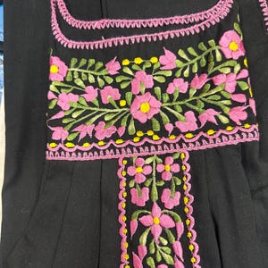 May include: Black garment with pink and yellow floral embroidery. The embroidery is featured around the neckline and down the front. The flowers are pink with yellow centers and green leaves. The garment has a square neckline.