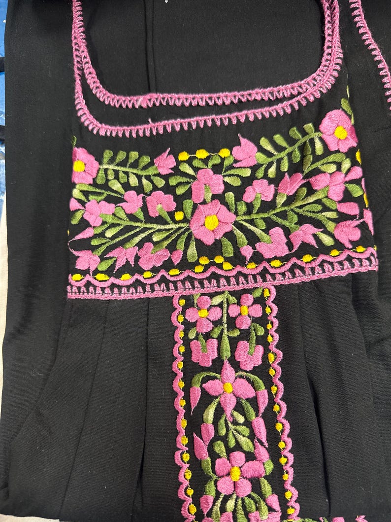 May include: Black garment with pink and yellow floral embroidery. The embroidery is featured around the neckline and down the front. The flowers are pink with yellow centers and green leaves. The garment has a square neckline.