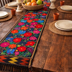 May include: A colourful, embroidered table runner featuring a floral design in red, blue, and pink. The runner is laid on a dark wooden table, with place settings, candles, and a basket of fruit. The runner has a black background and a fringed edge.