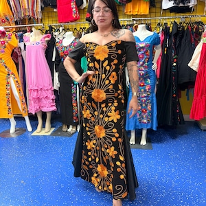 Pode incluir: Um vestido preto longo at&eacute; ao ch&atilde;o com ombros descobertos e bordados florais em laranja vibrante. A pe&ccedil;a tem mangas curtas, um corpete justo e uma saia rodada com padr&otilde;es de costura intrincados que se estendem da cintura para baixo.