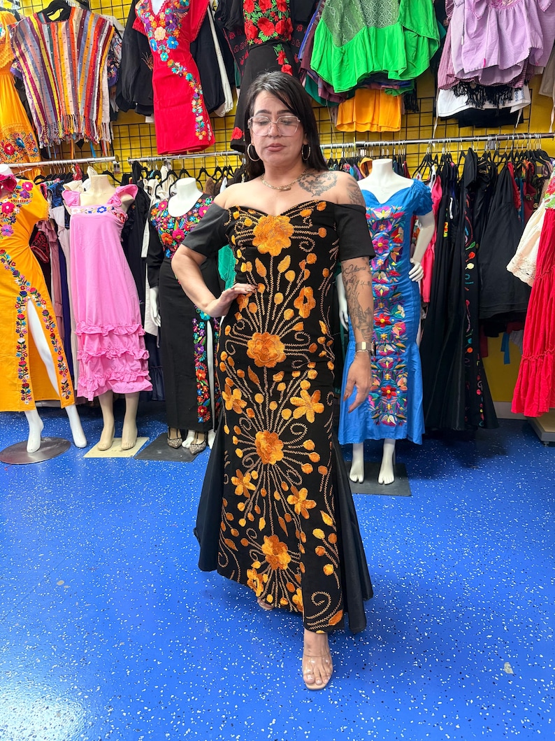 Pode incluir: Um vestido preto longo at&eacute; ao ch&atilde;o com ombros descobertos e bordados florais em laranja vibrante. A pe&ccedil;a tem mangas curtas, um corpete justo e uma saia rodada com padr&otilde;es de costura intrincados que se estendem da cintura para baixo.