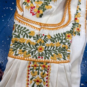 May include: Cream-colored embroidered garment featuring yellow and red floral designs with green leaves. The embroidery is outlined with orange stitching. The garment has a square neckline and a pleated skirt.