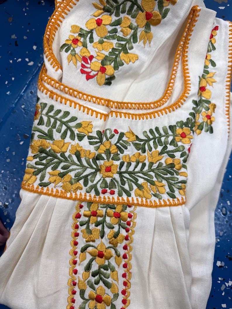 May include: Cream-colored embroidered garment featuring yellow and red floral designs with green leaves. The embroidery is outlined with orange stitching. The garment has a square neckline and a pleated skirt.