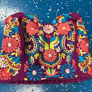 May include: A strapless purple corset top featuring vibrant, multi-colored floral embroidery. The design includes symmetrical patterns of coral, bright blue, and lime green flowers, along with two central bird figures stitched in blue and green thread.