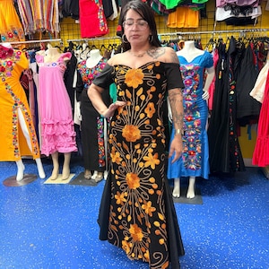 Pode incluir: Um vestido preto longo at&eacute; ao ch&atilde;o com ombros descobertos, mangas curtas e um padr&atilde;o vertical intrincado de flores cor de laranja bordadas e videiras que se estendem desde o corpete at&eacute; &agrave; bainha da pe&ccedil;a de vestu&aacute;rio.