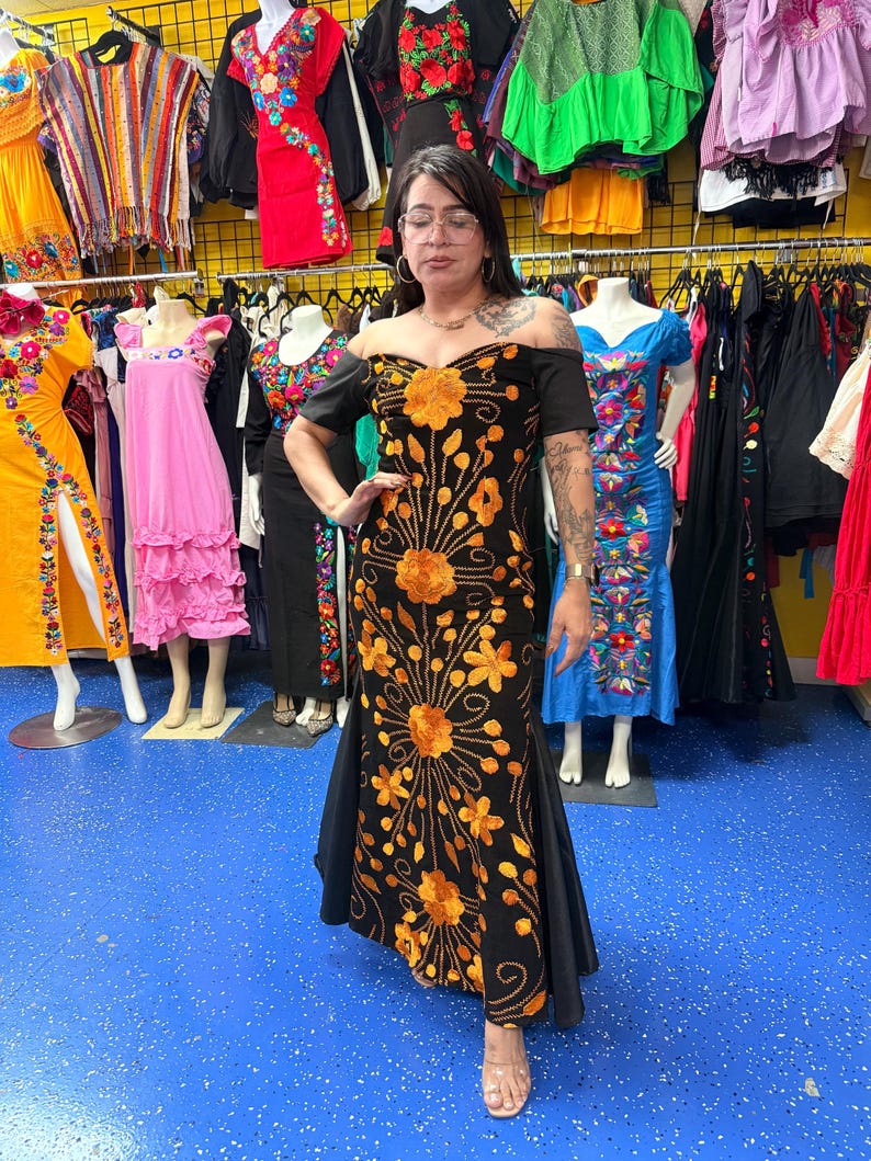 Pode incluir: Um vestido preto longo at&eacute; ao ch&atilde;o com ombros descobertos, mangas curtas e um padr&atilde;o vertical intrincado de flores cor de laranja bordadas e videiras que se estendem desde o corpete at&eacute; &agrave; bainha da pe&ccedil;a de vestu&aacute;rio.